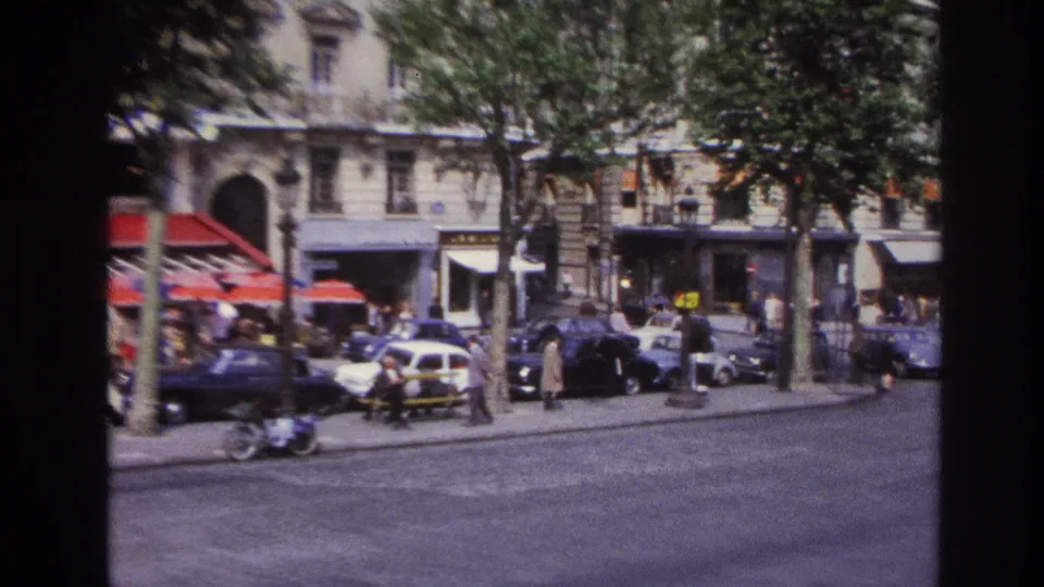 1967 Paris street frame with pedestrians near storefronts, parked classic cars, tree trunks, red awnings, and open roadway.