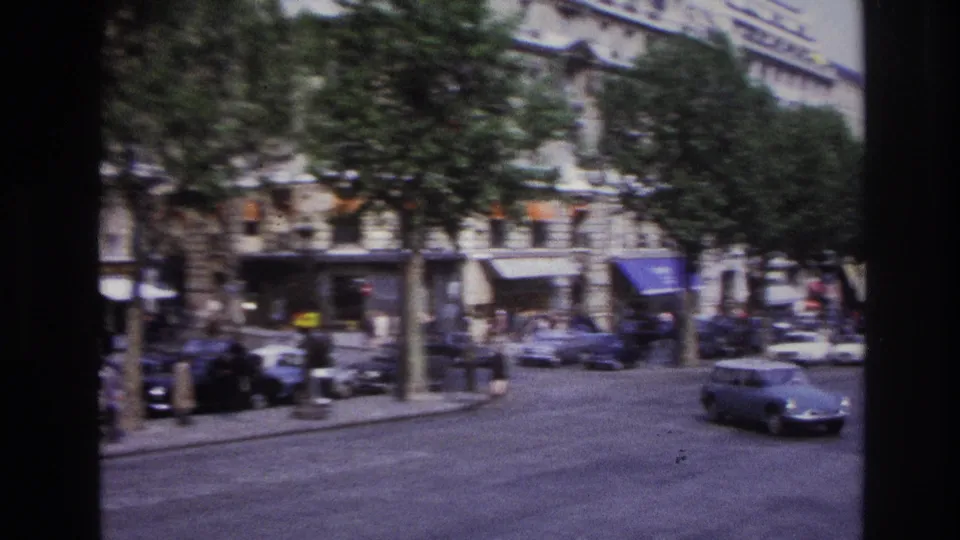 1967 Paris boulevard view showing storefronts, trees, pedestrians, parked cars, and a small car moving through the foreground.