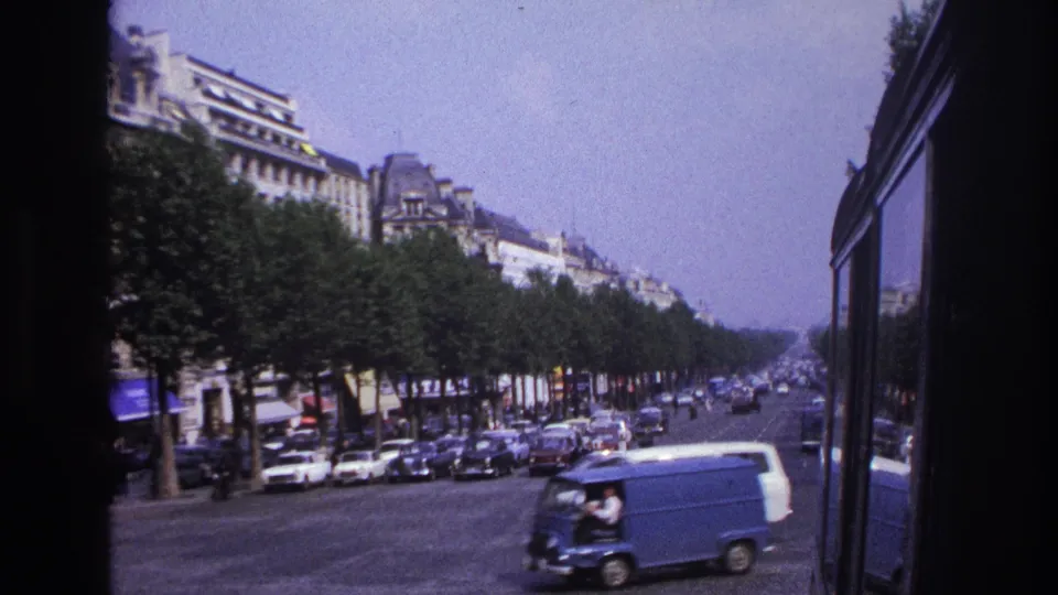 1967 Paris street view from a moving vehicle, with a blue van, parked cars, pedestrians, trees, and stone buildings visible.