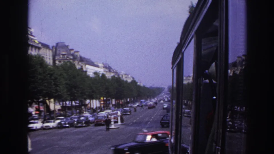1967 Paris moving-vehicle view showing the vehicle side at right, boulevard traffic, parked cars, street trees, and distant facades.