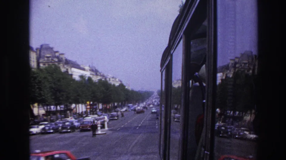 1967 Paris bus-window view down a broad tree-lined boulevard with mid-century cars, pedestrians, and multi-story buildings.