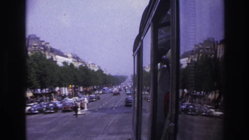 1967 Paris home-movie view from transit, with a long boulevard, mid-century cars, pedestrians, trees, and building facades.