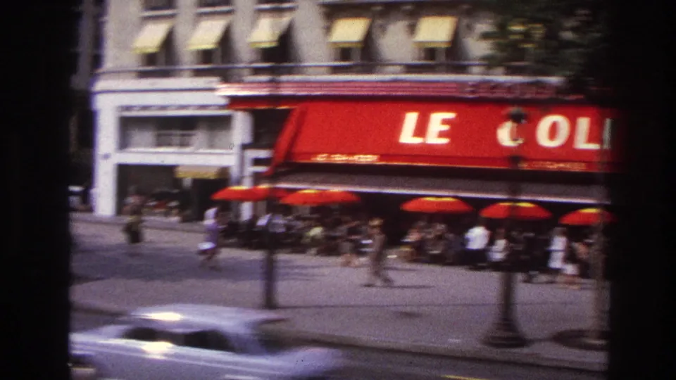 1967 Paris street-level bus view of a cafe exterior with a red LE COLISEE awning, umbrellas, seated patrons, and passing traffic.