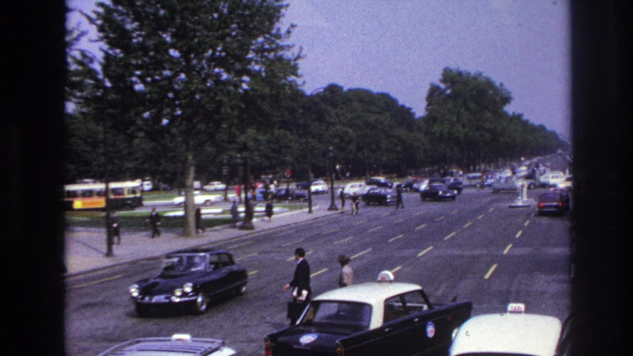 1967 Paris street view from a moving vehicle, with pedestrians crossing a broad boulevard among taxis, cars, and a yellow bus.