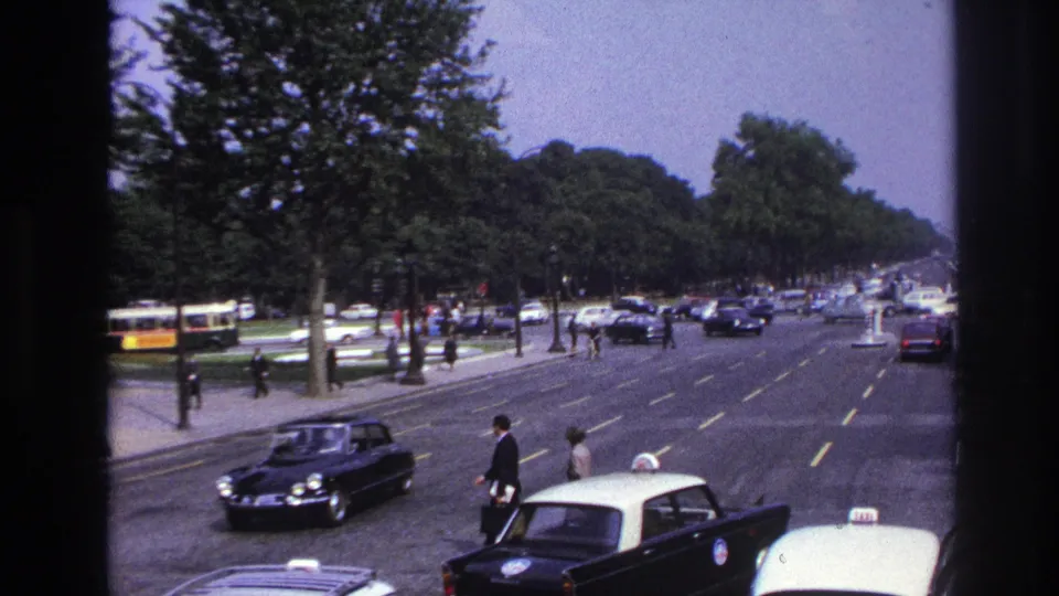 1967 Paris street view from a moving vehicle, with pedestrians crossing a broad boulevard among taxis, cars, and a yellow bus.