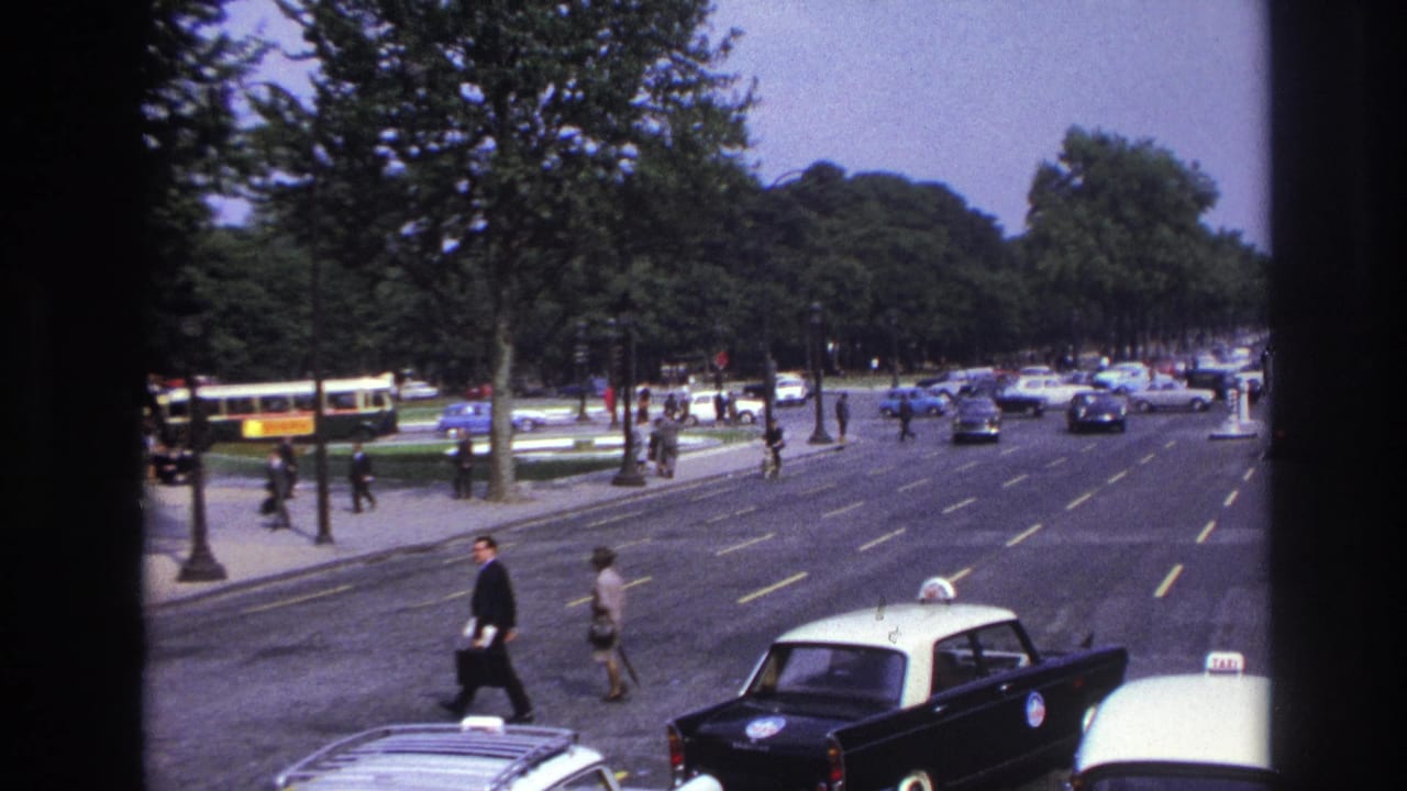 Two pedestrians cross near taxis on a broad boulevard with cars and buses in the distance.