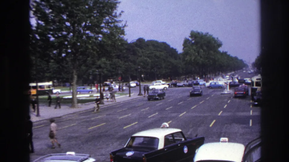 Home-movie view of a 1967 Paris boulevard with a pedestrian crossing, taxis in the foreground, and classic traffic beyond.