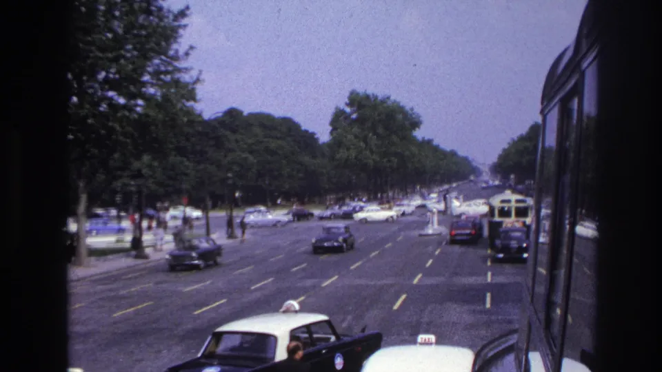 1967 Paris traffic view from a moving vehicle, showing taxis, classic cars, a bus edge, and pedestrians along a tree-lined boulevard.