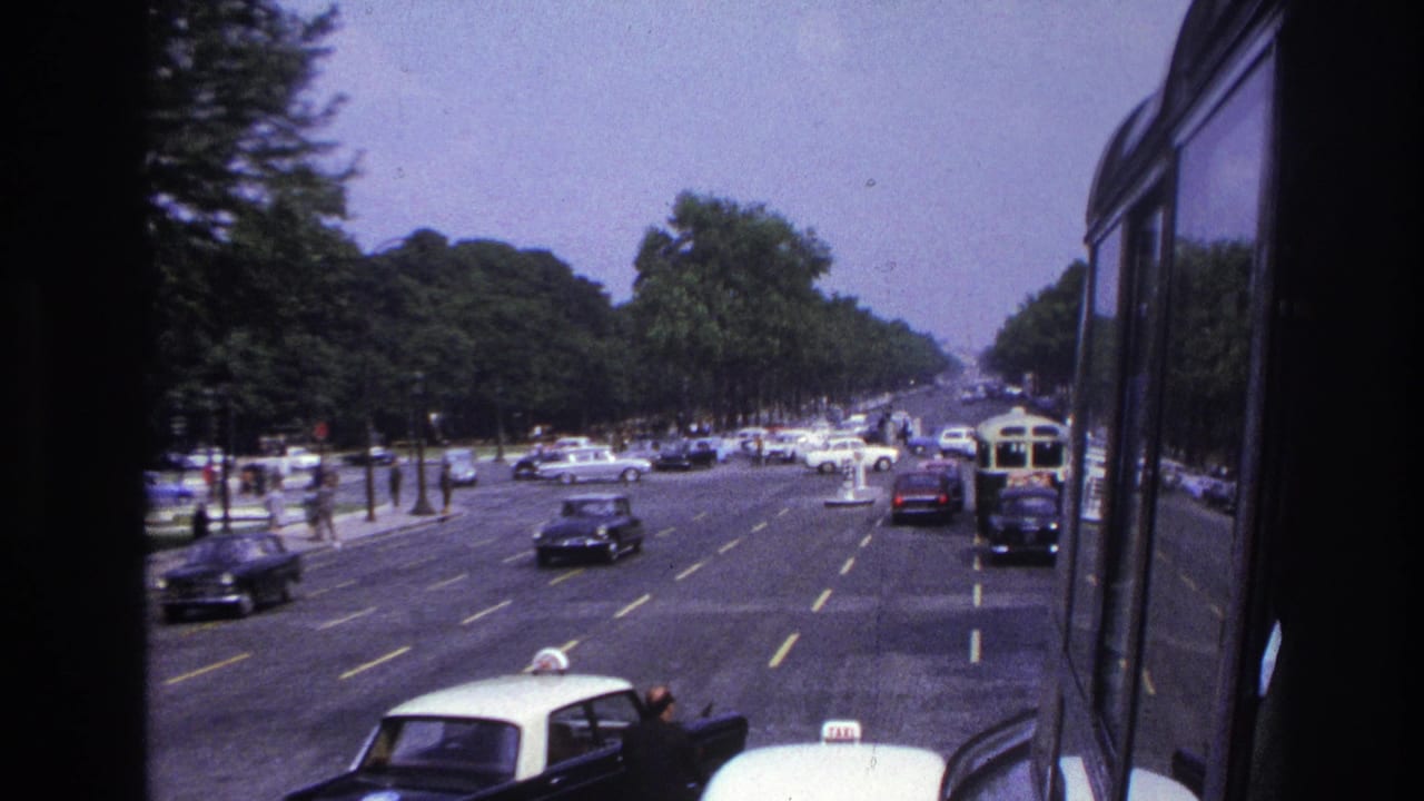 1967 Paris street traffic viewed from a moving vehicle, with cars, a bus, trees, and a dark window edge in frame.