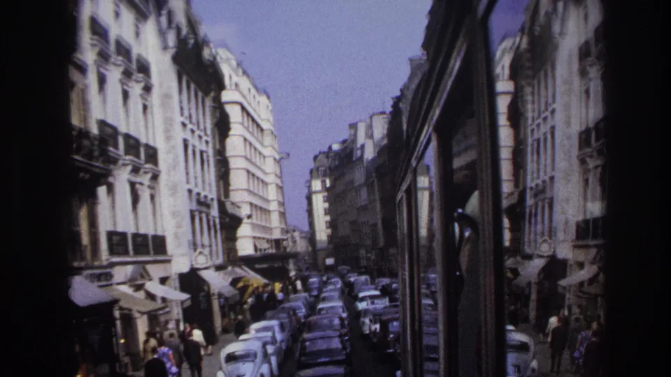 1967 Paris street scene from a moving vehicle, showing parked cars, pedestrians, awnings, and multi-story buildings.