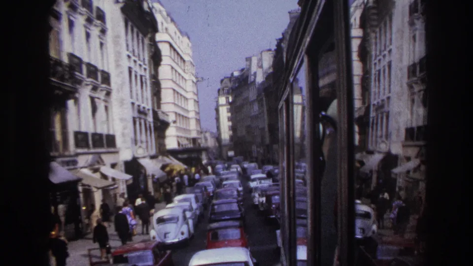 1967 Paris street view with dense parked cars, pedestrians on sidewalks, shop awnings, and the side of the filming vehicle.