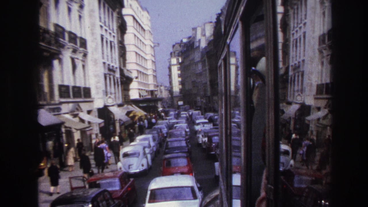 1967 Paris street view from a moving bus, with compact cars, pedestrians, shop awnings, and multi-story building facades.