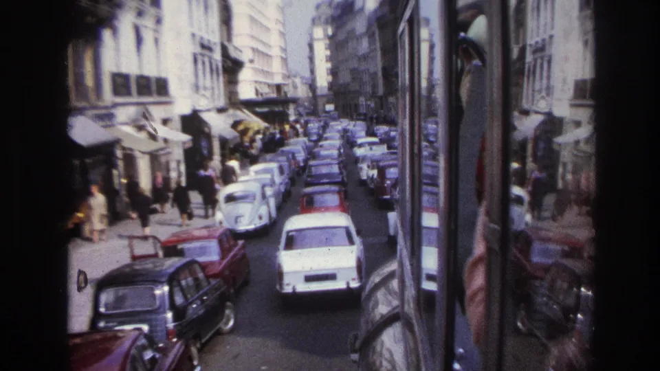 1967 Paris street scene from a bus, showing a white Volkswagen Beetle, surrounding cars, pedestrians, and shopfront awnings.