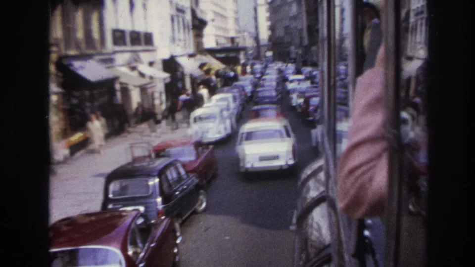 Late-1960s Paris bus-window view with compact cars, pedestrians, shop awnings, and a passenger or passerby partly visible at right.