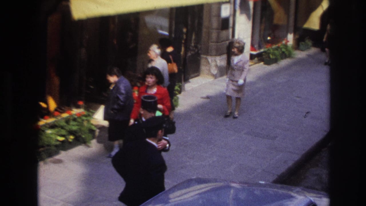Paris sidewalk scene with pedestrians in formal clothing, flower planters by a storefront, and a uniformed hotel or service worker seen from above.