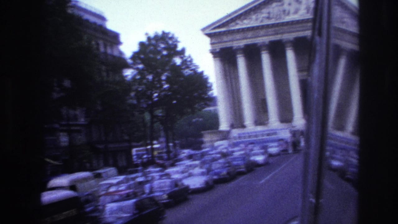 1967 Paris home-movie frame showing parked cars, trees, and a large neoclassical building seen from inside a moving vehicle.