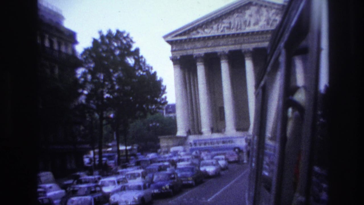Street scene with traffic, trees, and a neoclassical building viewed through a vehicle window.