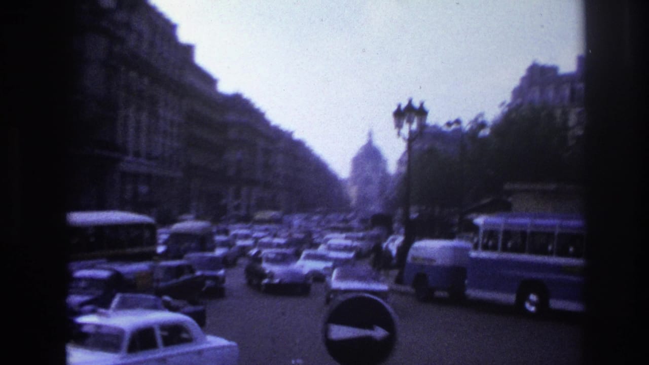 1967 Paris home-movie view of boulevard traffic, buses, tall buildings, street lamps, and a distant arch-like monument.