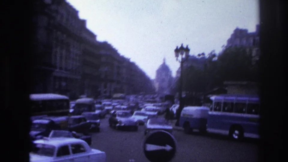 1967 Paris home-movie view of boulevard traffic, buses, tall buildings, street lamps, and a distant arch-like monument.