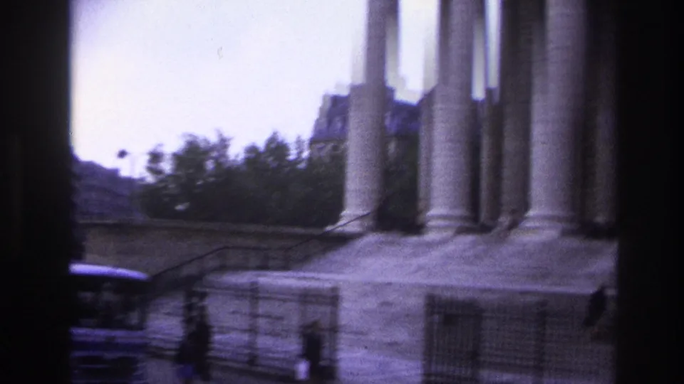 Handheld 1967 Paris view of a monumental columned building facade with steps, railings, trees, and passing street edge.