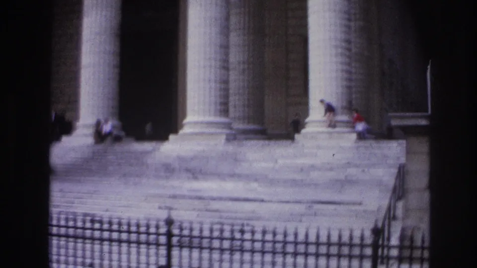 1967 Paris home-movie frame showing a classical colonnaded building, broad steps, iron fence, and small figures near the entrance.