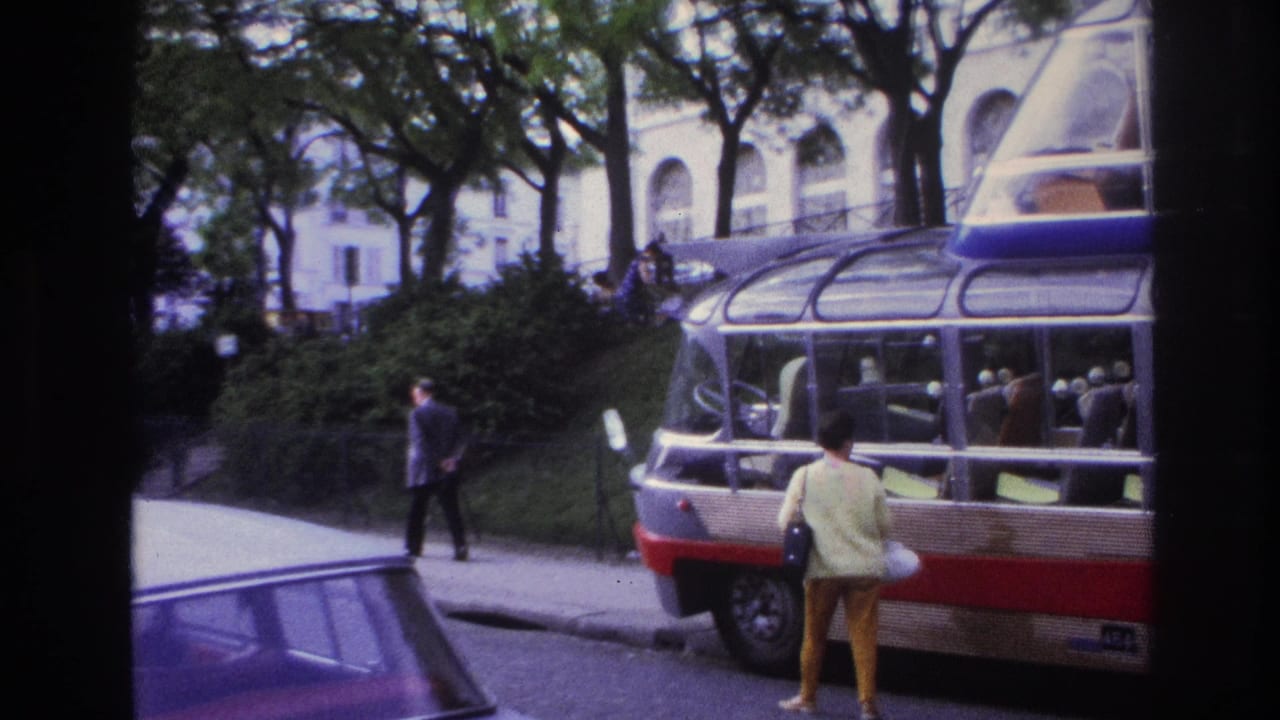1967 Paris home-movie frame showing a woman posing near a glass-roof sightseeing bus, with trees, pedestrians, and a pale arched building behind.