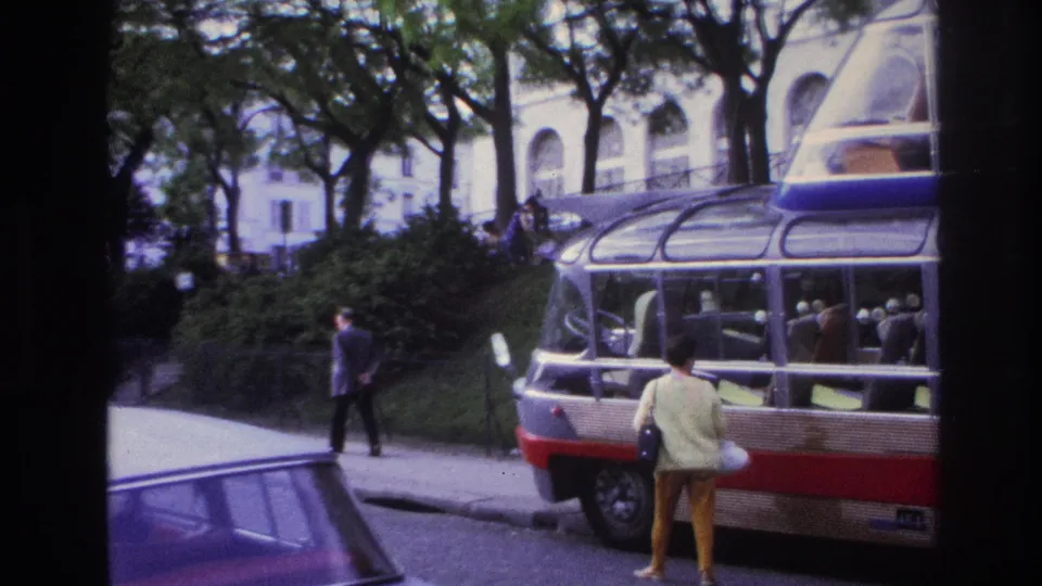 1967 Paris home-movie frame showing a woman posing near a glass-roof sightseeing bus, with trees, pedestrians, and a pale arched building behind.