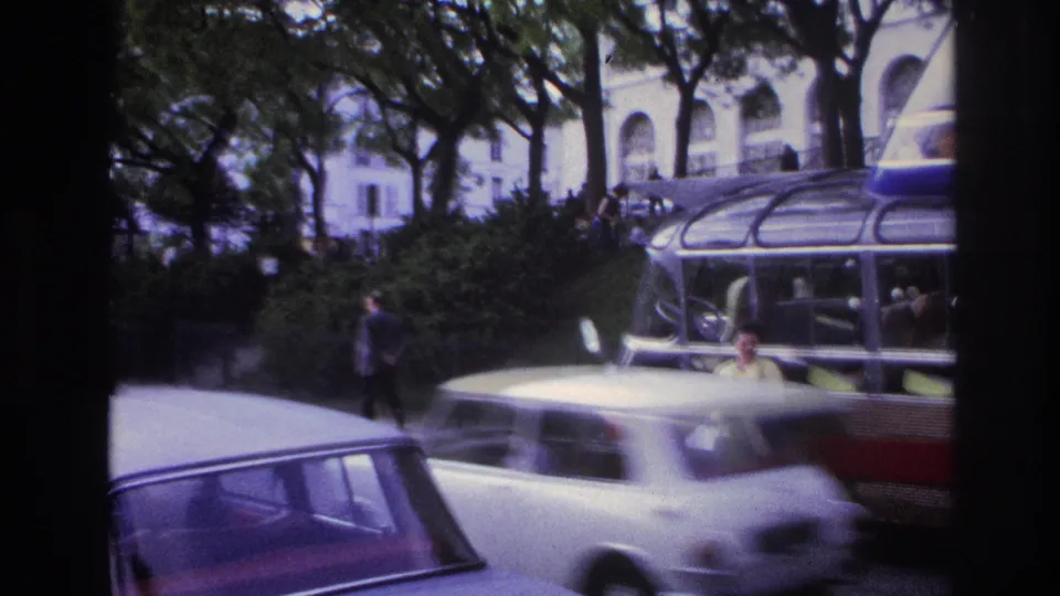 A white car blurs past a paused sightseeing bus in 1967 Paris as a woman stands near the street and trees fill the background.