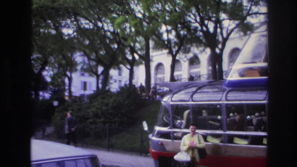 The woman in a yellow top faces the camera beside the glass-roof bus, with a pedestrian, trees, and a pale arched building visible behind.