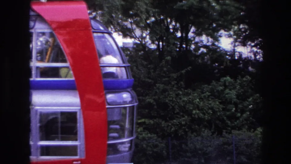 1967 Paris 8mm frame showing a cropped glass-sided structure, red support, dark foreground edge, and greenery.
