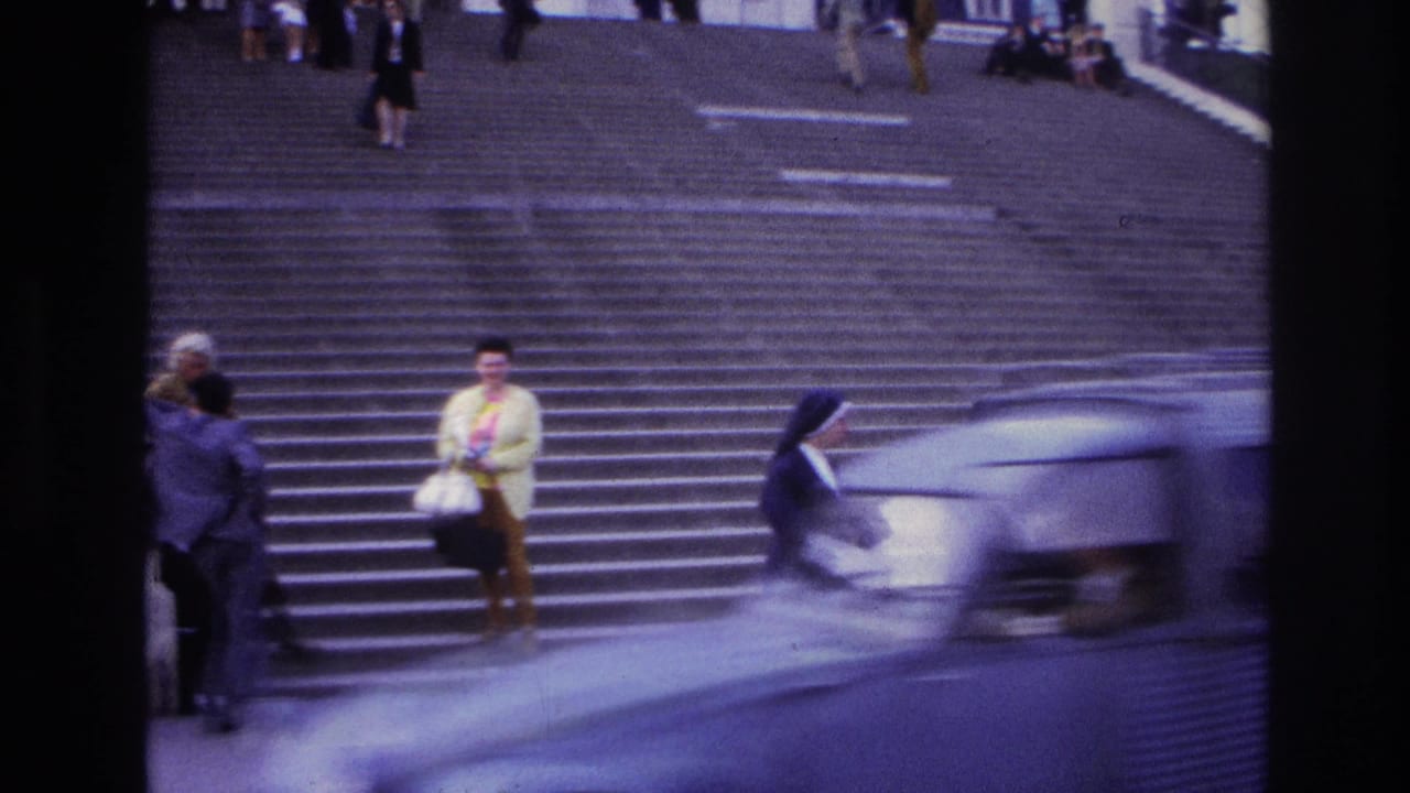 A car blurs across the foreground while pedestrians stand and walk on the steps behind it.