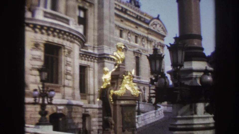 1967 Paris home-movie frame showing an ornate facade with gilded sculptural details, street lamps, steps and a column.