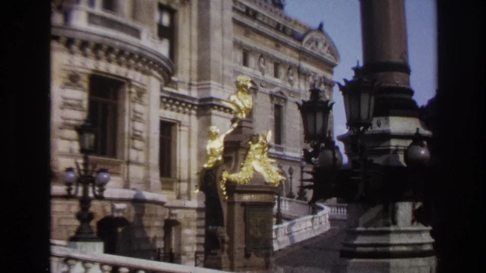 1967 Paris home-movie frame showing a grand stone facade with gilded sculpture, dark lampposts, and a curved balustrade.