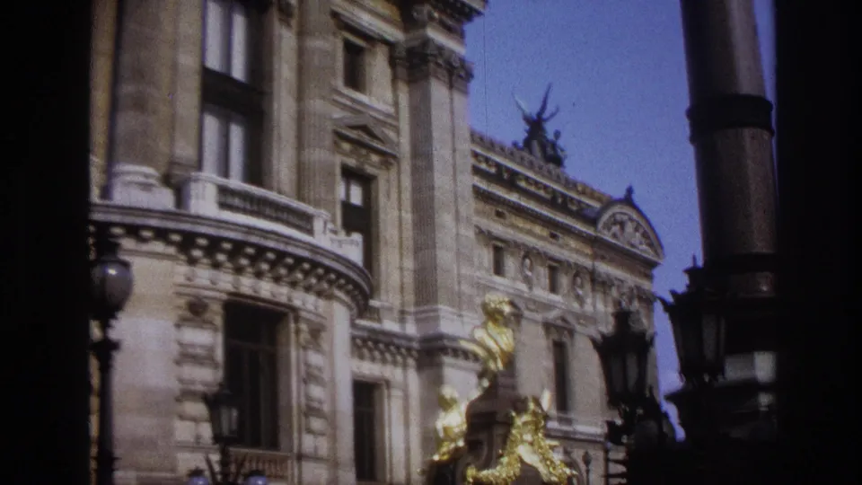 1967 Paris exterior frame with a grand stone facade, gilded sculptural detail, ornate roofline, and foreground lamps.