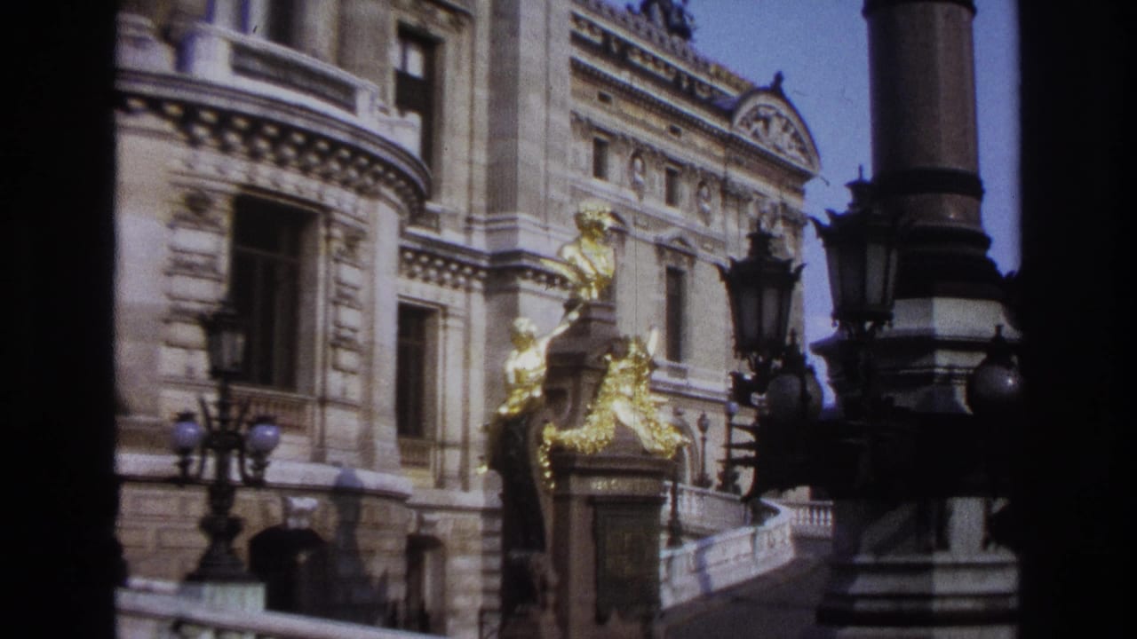 Stone facade, gold statue, columns, and lamps seen through dark vertical foreground shapes.