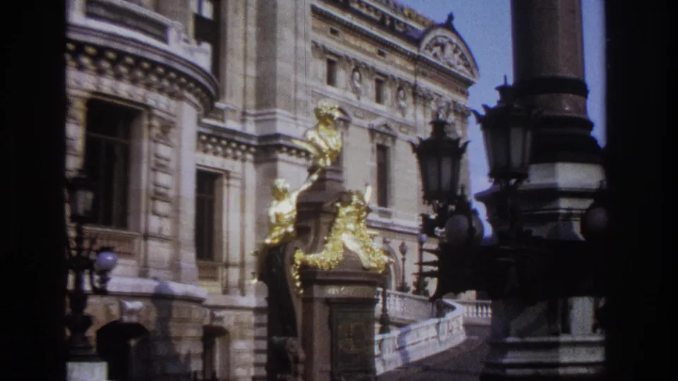 1967 Paris 8mm frame showing gold figures on a pedestal, carved facade details, windows, railings, and black lamp posts.