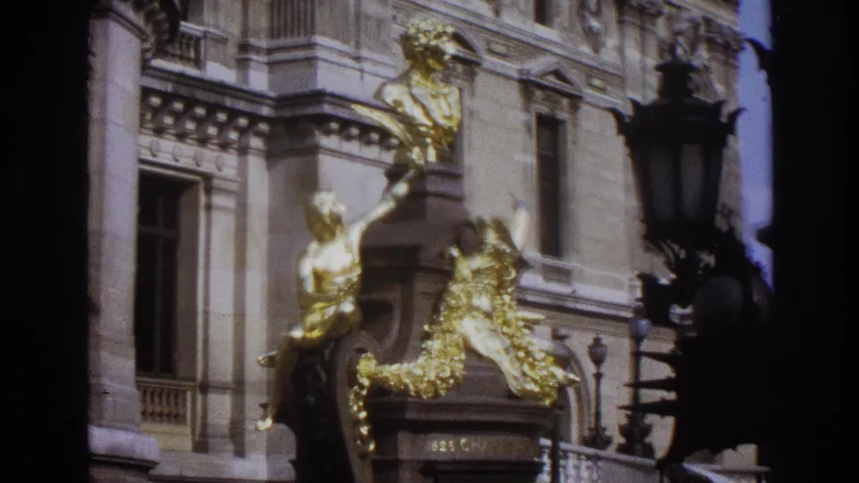 1967 Paris home-movie frame showing gilded sculptural figures on a grand stone building facade.