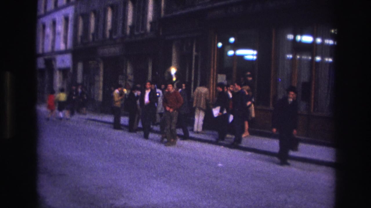 1967 Paris home-movie frame showing adults gathered along a sidewalk near shop windows as a man moves through the street.