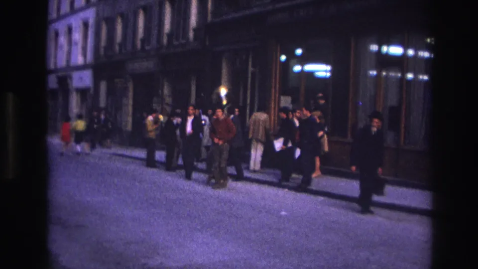 1967 Paris home-movie frame showing adults gathered along a sidewalk near shop windows as a man moves through the street.