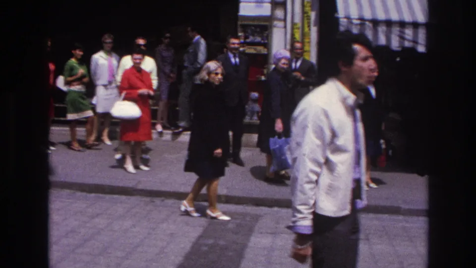 1967 Paris home-movie frame showing the man in a white jacket walking past women with handbags and suited men near a shopfront.