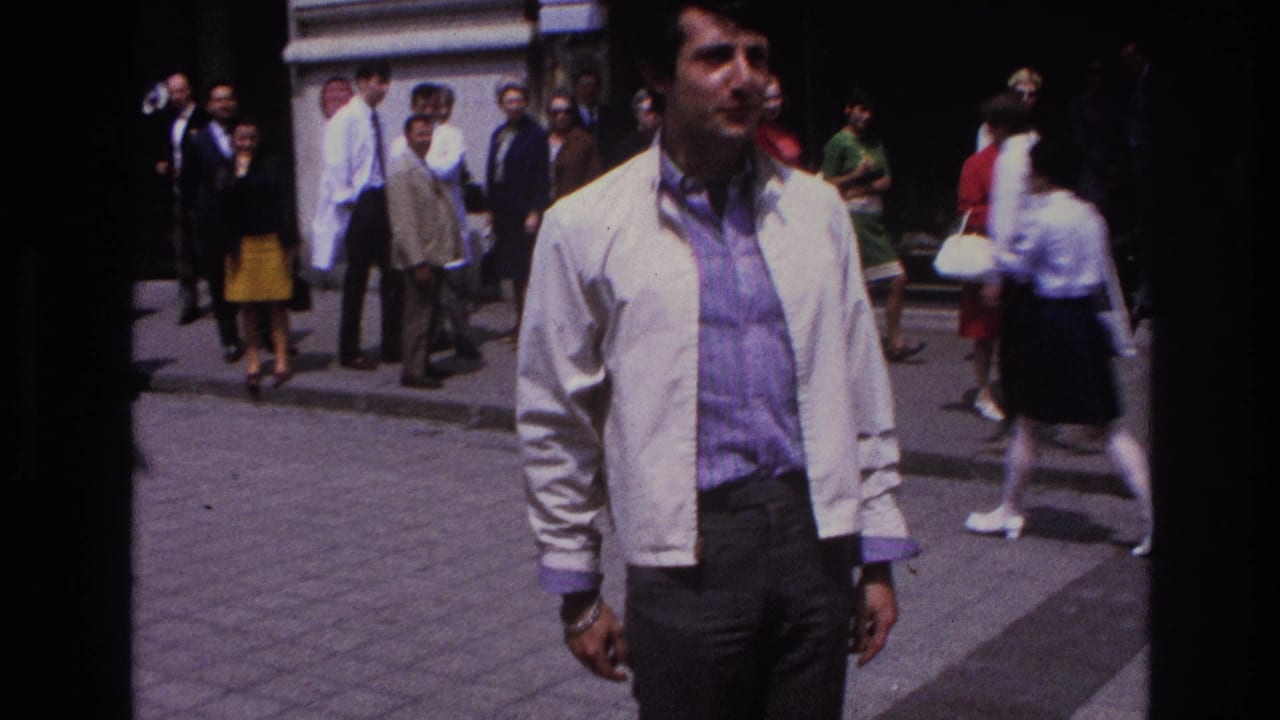 Man in a white jacket stands in the foreground with pedestrians gathered behind him.