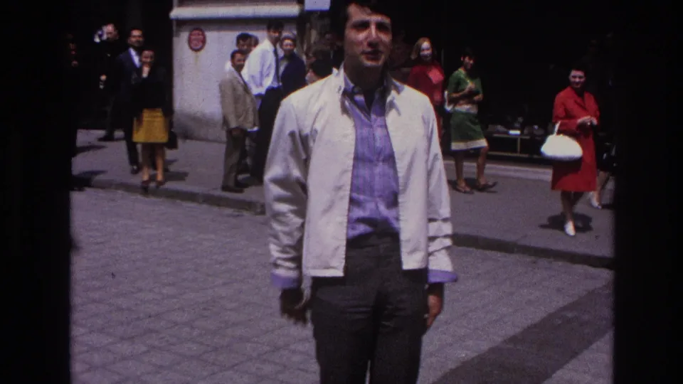 The man stands near center frame in 1967 Paris street footage, with pedestrians in period clothing and a shaded storefront area behind him.