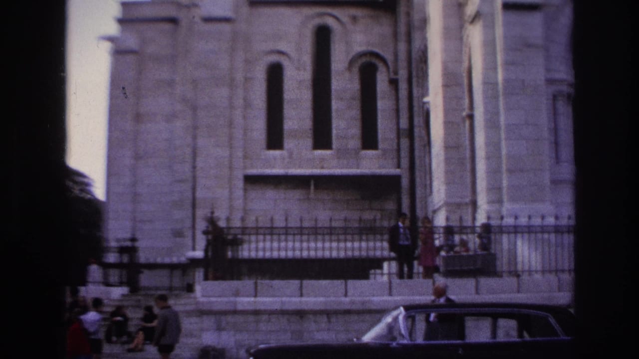 1967 Paris home-movie frame showing a large stone religious facade, iron railings, people near the sidewalk, and a dark sedan passing or parked below.