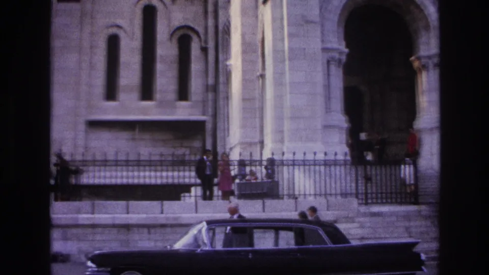 1967 Paris frame with a dark mid-century sedan along the curb, people by an iron fence, and a stone church facade with tall arched windows behind them.