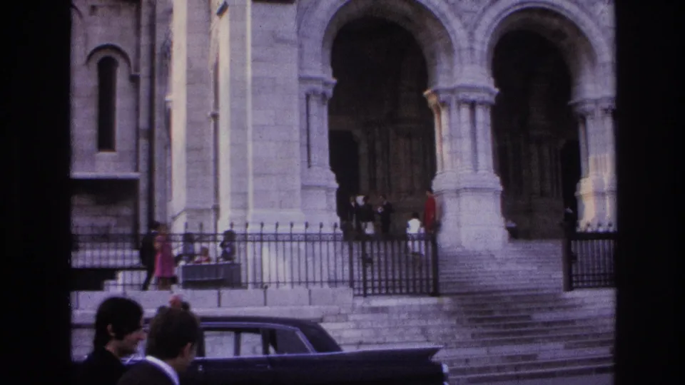 1967 Paris home-movie view of a large arched church entrance with steps, iron fencing, pedestrians, and a dark car partly visible at the bottom.