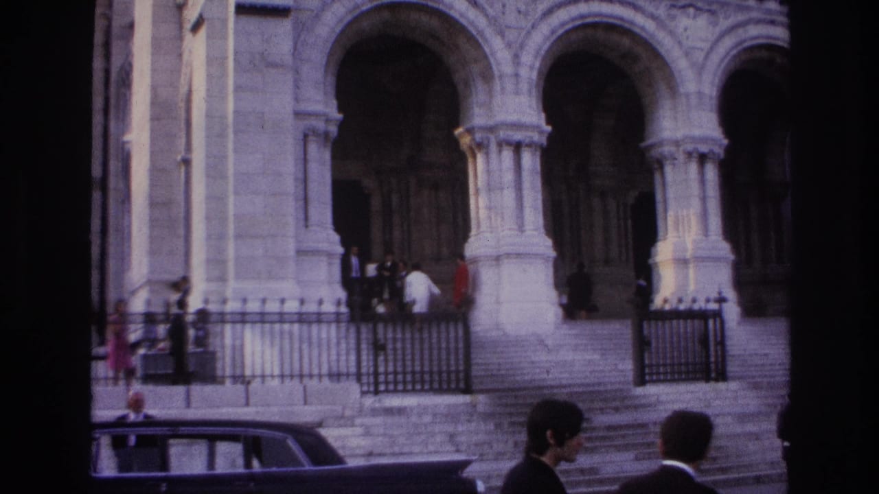 1967 Paris home-movie frame showing a grand stone entrance with arches, steps, iron railings, pedestrians, and a dark car.