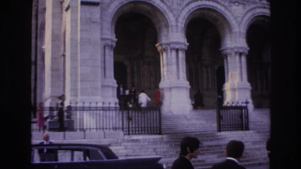 1967 Paris home-movie frame showing a grand stone entrance with arches, steps, iron railings, pedestrians, and a dark car.