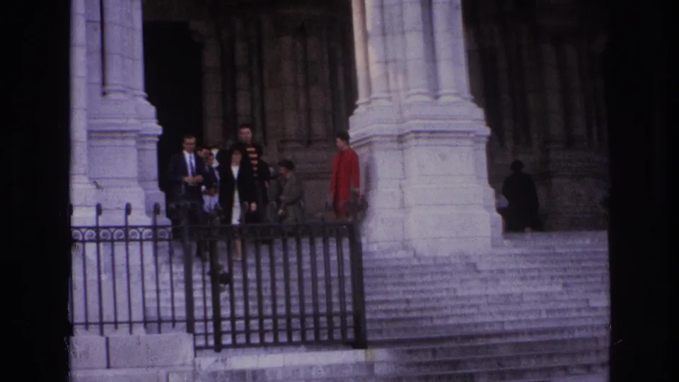 1967 Paris home-movie still with suited visitors, a red coat, pale stone columns, steps, and black railings.