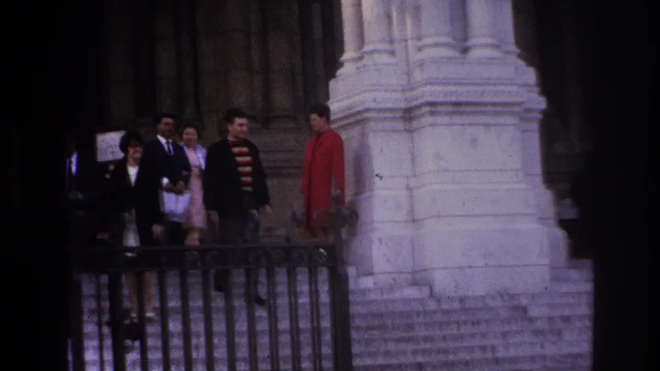 1967 Paris frame showing visitors descending monumental steps, including a person in a red coat, beside pale columns and railings.