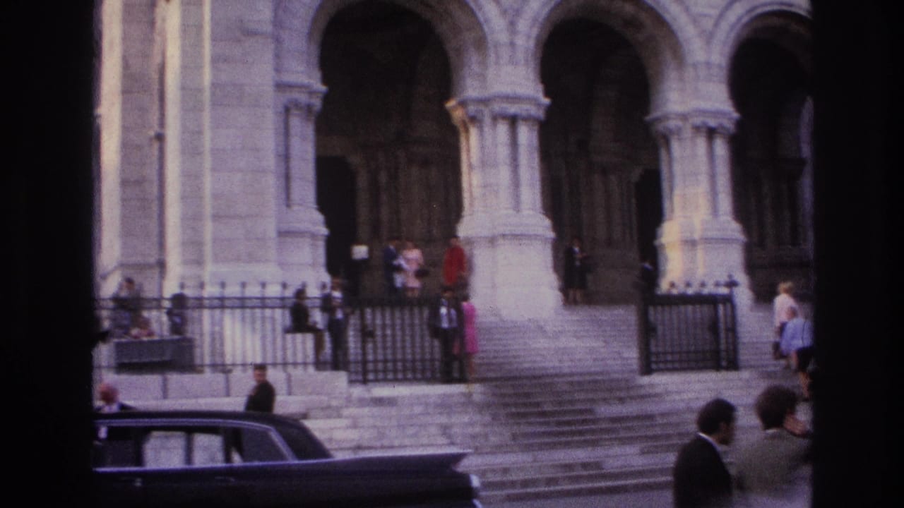 1967 Paris home-movie frame of a large stone religious facade with arched portals, steps, visitors, fencing, and a dark car nearby.